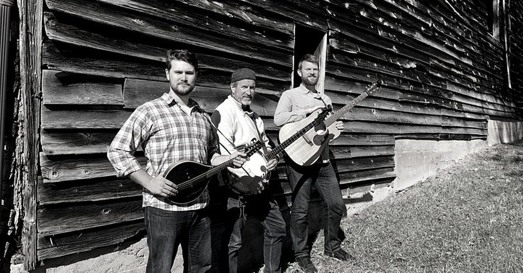 Three-piece string band with banjo and guitars standing against a weathered barn