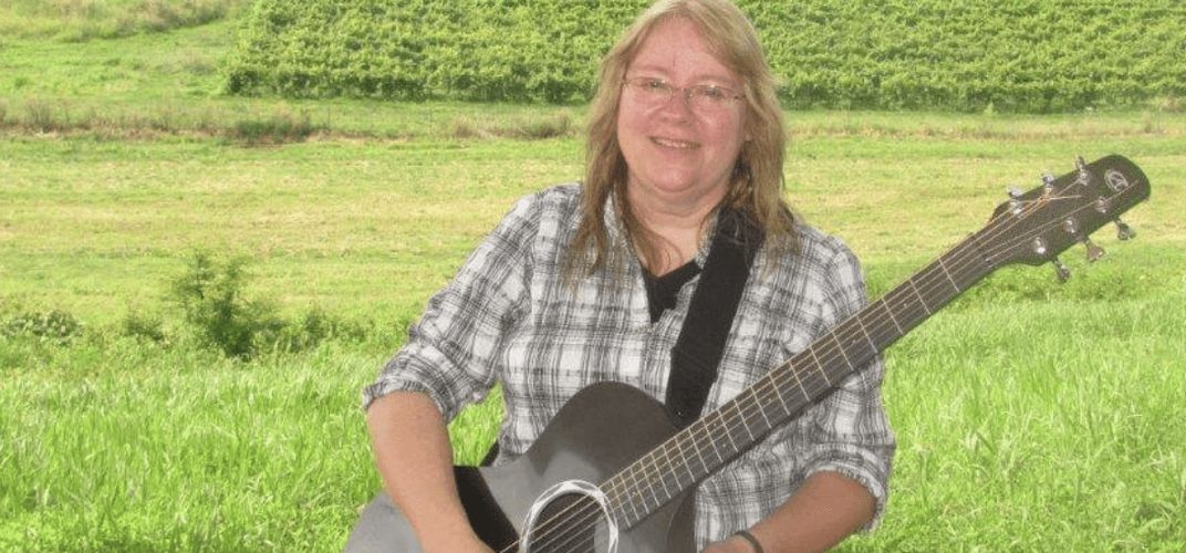 Singer-songwriter Sue Harlow holding an acoustic guitar in an open green field