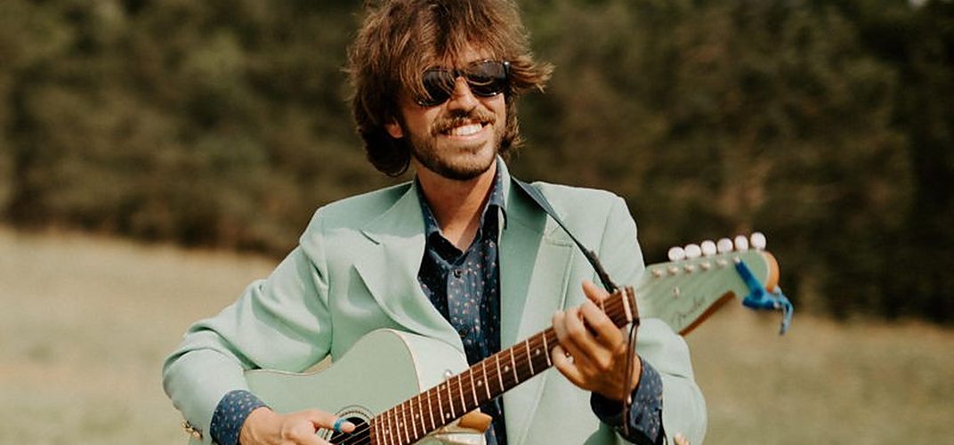 Grayson Harlow in a pale green suit playing electric guitar in an open field